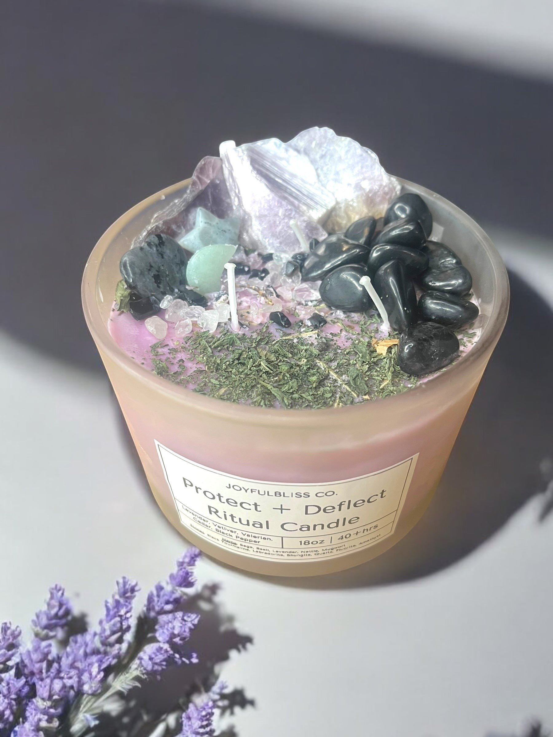 JoyfulBliss Co. Protect + Deflect soy candle topped with black tourmaline, amethyst, and herbs in a frosted glass jar.