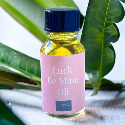 JoyfulBliss Co. Luck Be Mine ritual oil in a glass bottle with a pink label, set against green leaves.