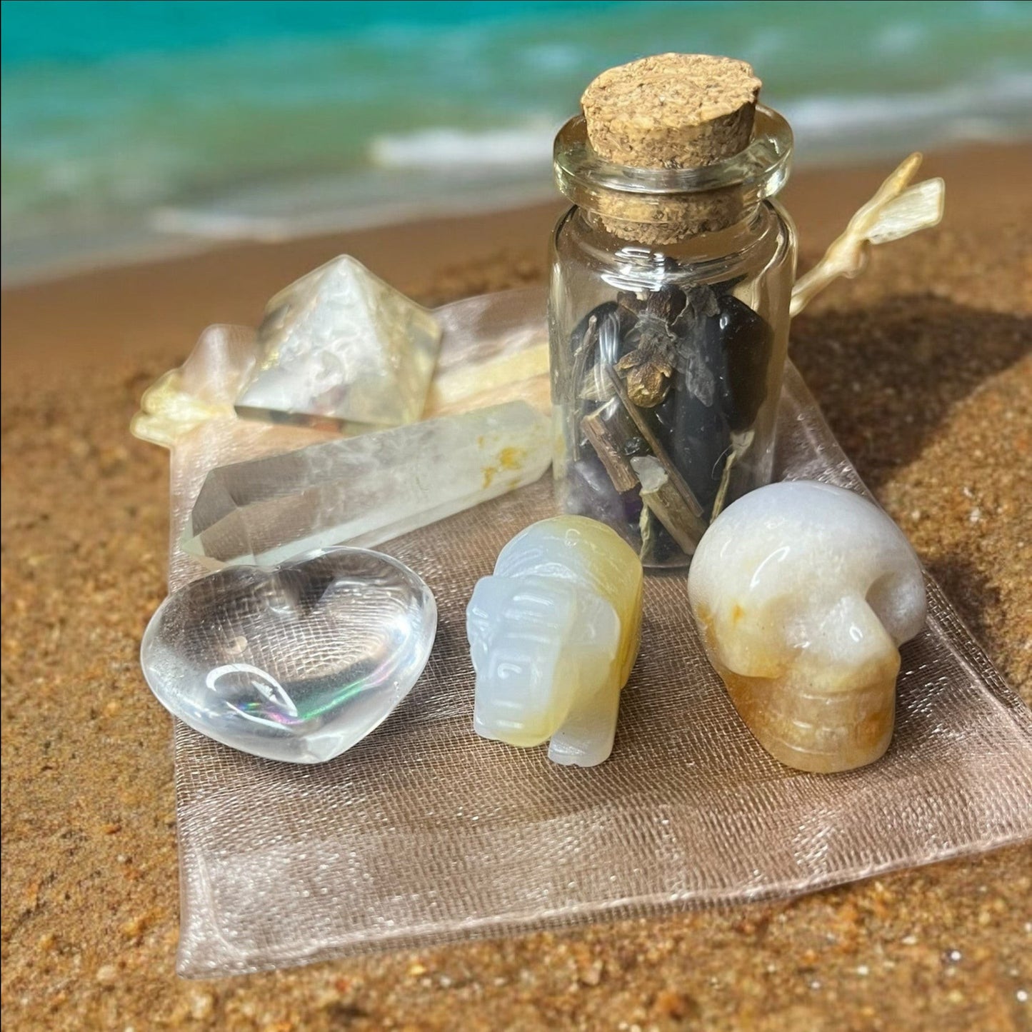 JoyfulBliss Co. Manifesting Mojo Bag with crystals, spell jar, and heart, elephant, and skull carvings on a beach.