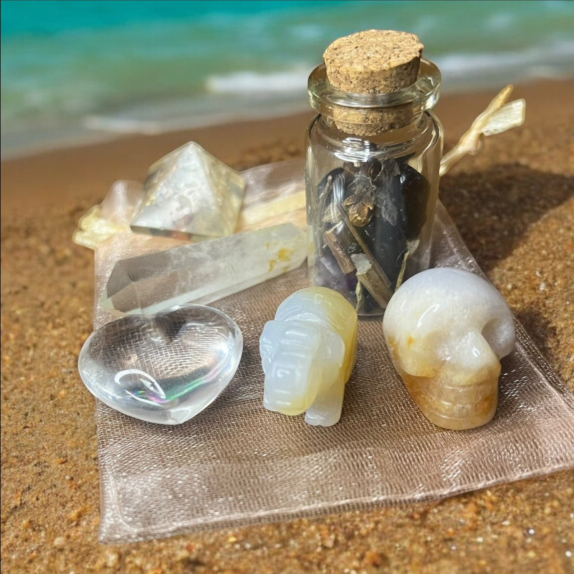 JoyfulBliss Co. Manifesting Mojo Bag with crystals, spell jar, and heart, elephant, and skull carvings on a beach.