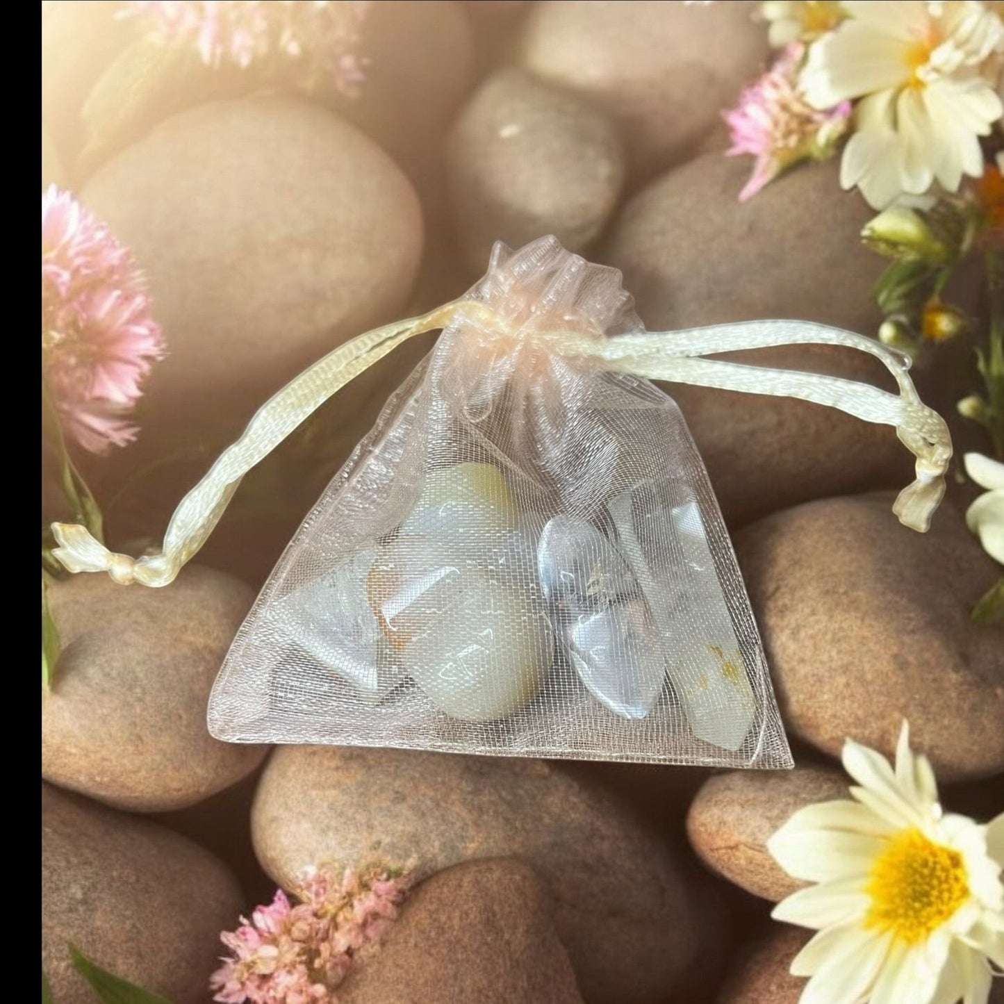 JoyfulBliss Co. Manifesting Mojo Bag with clear quartz crystals in a sheer organza pouch on a bed of stones and flowers