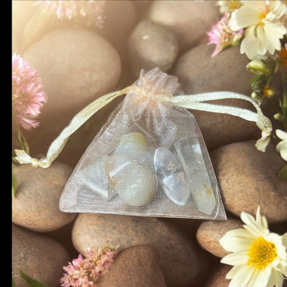 JoyfulBliss Co. Manifesting Mojo Bag with clear quartz crystals in a sheer organza pouch on a bed of stones and flowers