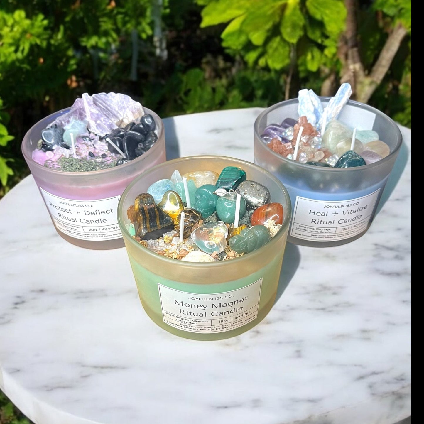 JoyfulBliss Co. set of three ritual candles topped with healing crystals and herbs on a marble table.